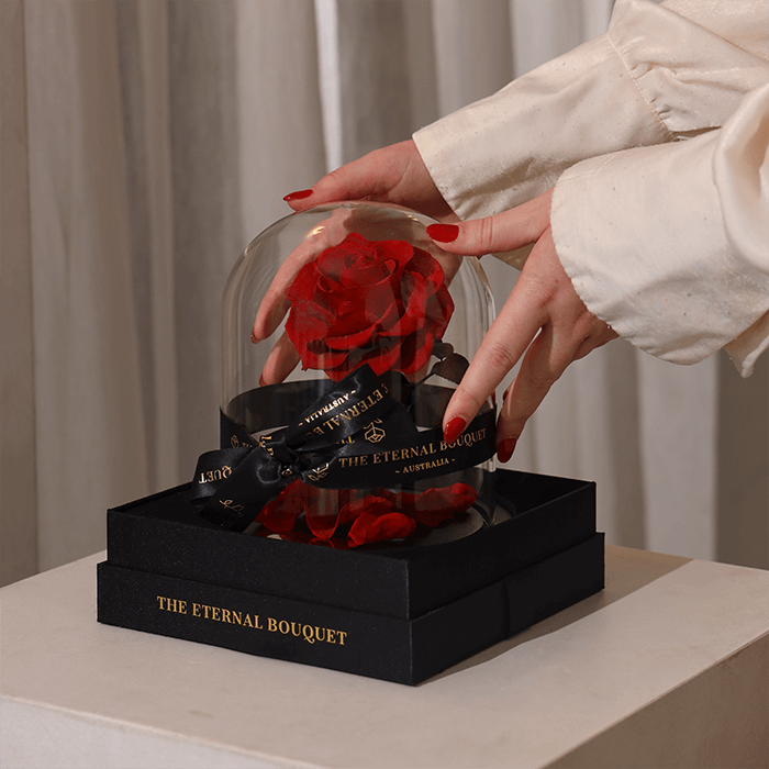 Hands carefully placing an everlasting red rose encased in a glass dome onto a black gift box with gold "The Eternal Bouquet" branding—an elegant and thoughtful luxury gift perfect for romantic occasions.