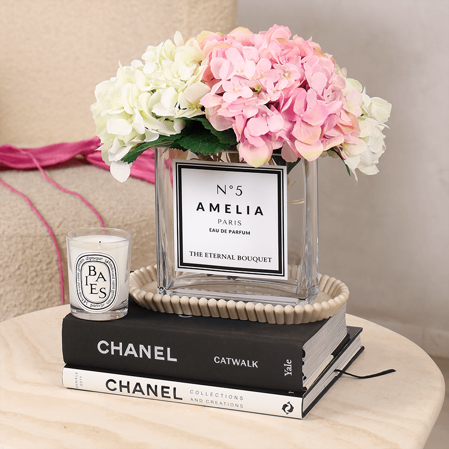 A luxurious floral arrangement by The Eternal Bouquet, featuring elegant white and soft pink hydrangeas displayed in a clear, square glass vase. The vase has a sleek white personalised label with black text, styled like a designer perfume bottle, reading "N°5 CHANEL PARIS EAU DE PARFUM" and "THE ETERNAL BOUQUET" at the bottom. The arrangement sits on a white surface with soft, sheer curtains in the background, creating an elegant and sophisticated presentation.