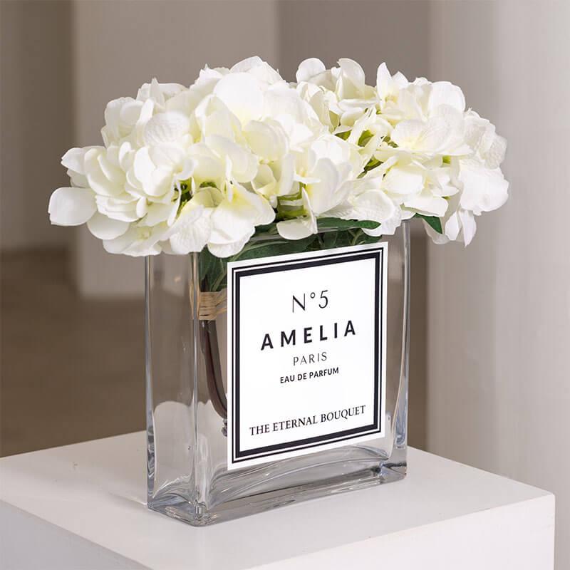 A personalised glass vase from The Eternal Bouquet filled with elegant white everlasting hydrangeas. The label reads 'N°5 Amelia Paris Eau de Parfum,' offering a timeless and sophisticated gift perfect for any occasion.