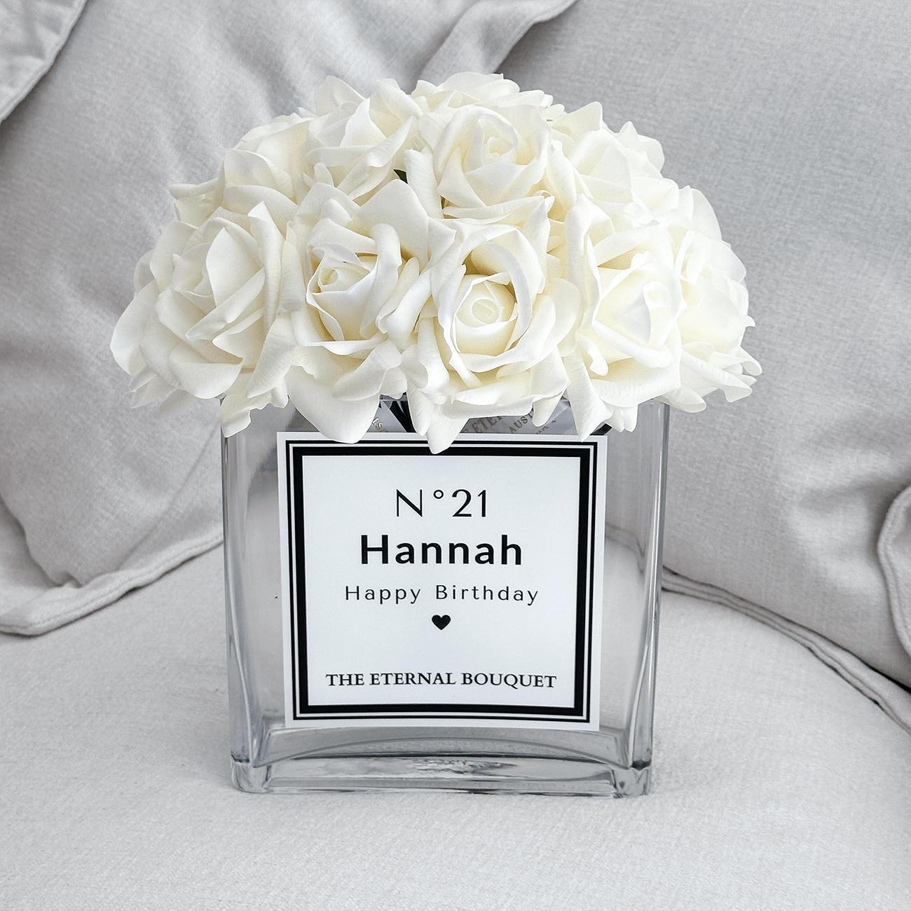 White silk roses arranged in The Eternal Bouquet Personalised glass vase on an off-white sofa with two cushions.