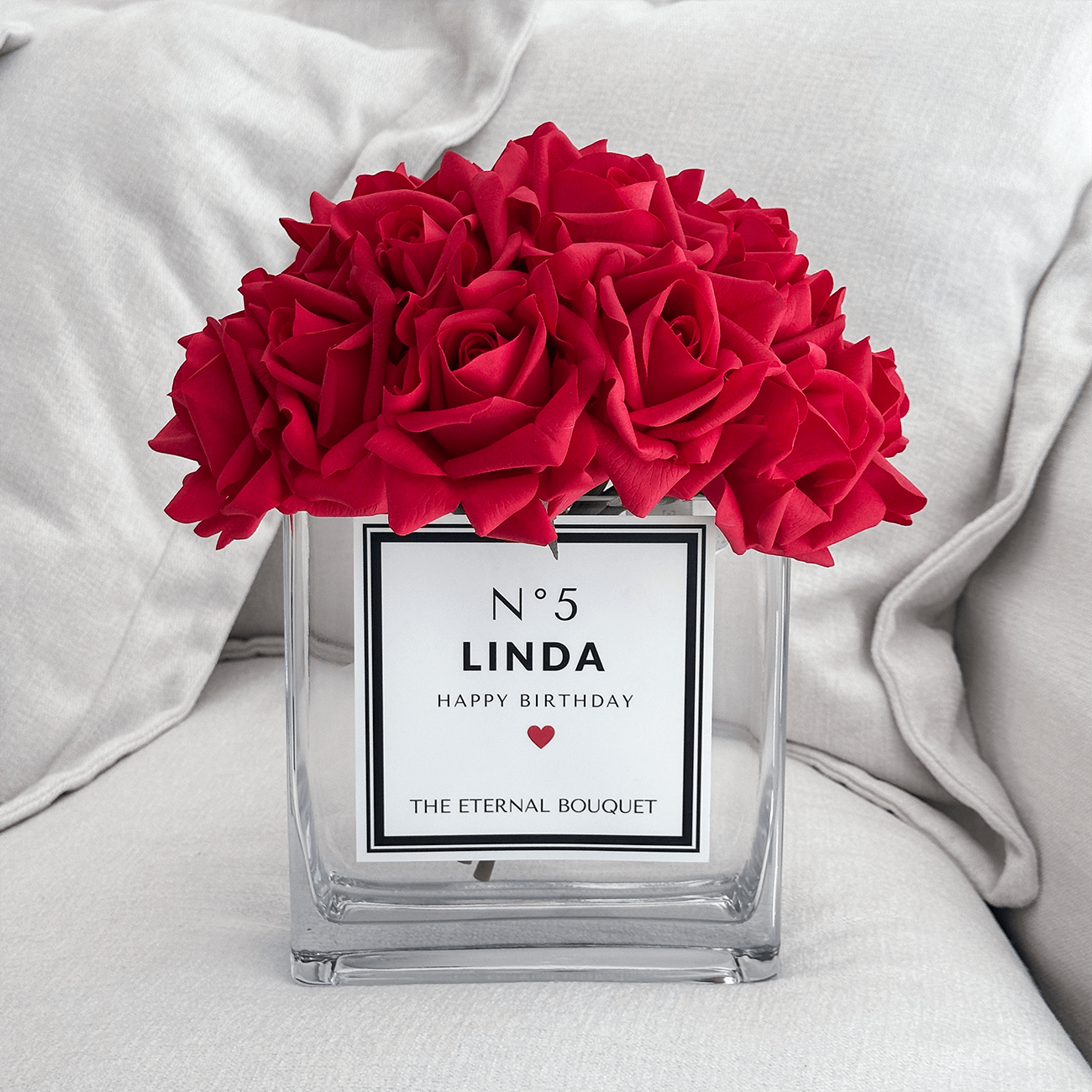 The Eternal Bouquet Personalised glass vase arranged with red silk roses on an off-white sofa.