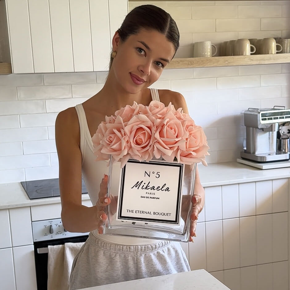 Young brunette woman holding a personalised glass vase with blush roses in the kitchen.