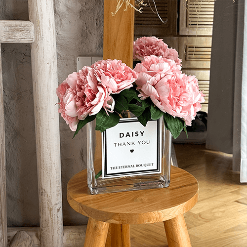 A bouquet of pink peonies with lush green leaves arranged in a clear glass vase with a minimalist label. The vase sits on a round wooden stool in a cozy, modern interior with neutral tones, a wooden mirror, and decorative elements in the background.