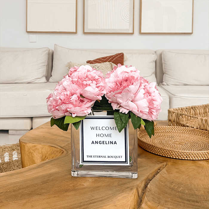 A luxurious bouquet of soft pink peonies with lush green leaves, elegantly arranged in a clear glass vase. The vase features a sleek, modern label.