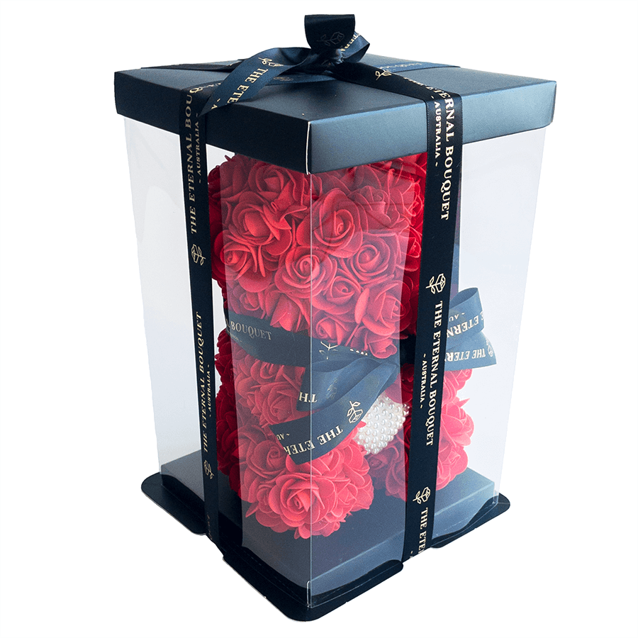 An everlasting luxury red rose teddy bear from The Eternal Bouquet, beautifully displayed in a clear gift box with black and gold branded ribbon. Featuring a pearl heart embellishment at the center, this elegant floral arrangement offers a premium unboxing experience—ideal for romantic gestures, proposals, or celebrating someone special in a truly unforgettable way