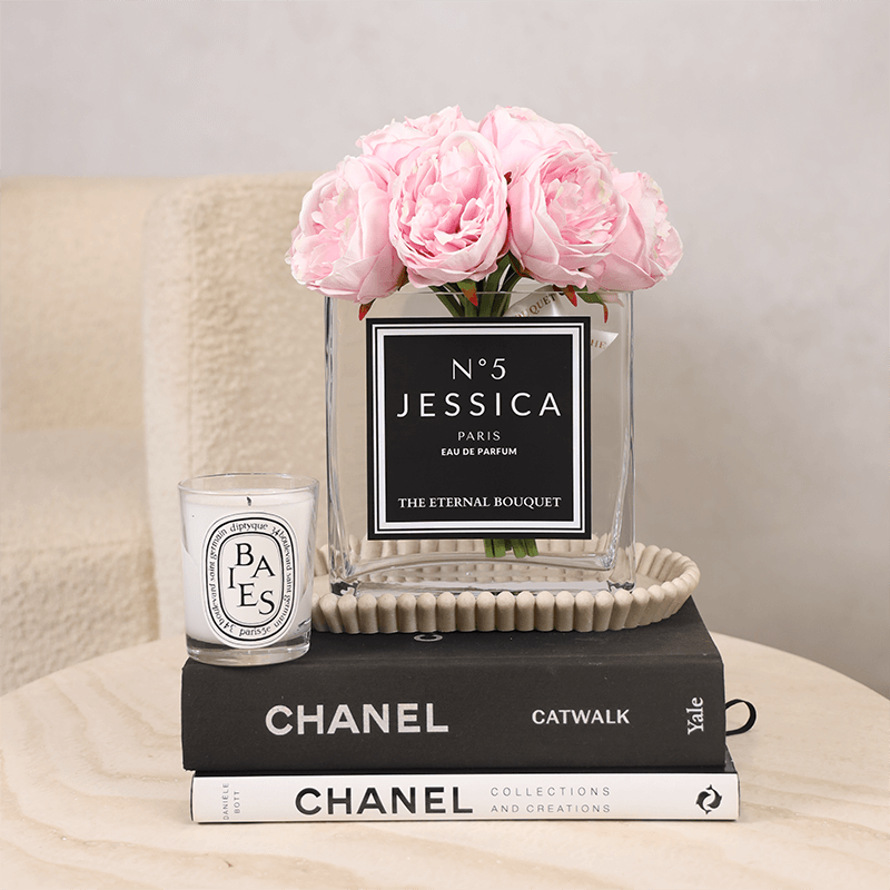 A luxurious floral arrangement from The Eternal Bouquet featuring soft pink peonies displayed in a clear, square glass vase. The vase is personalized with a sleek white label. Surrounding the main arrangement are additional personalized bouquets, creating an elegant and romantic setting. Perfect as gifts for your partners, or to commemorate meanignful memories.