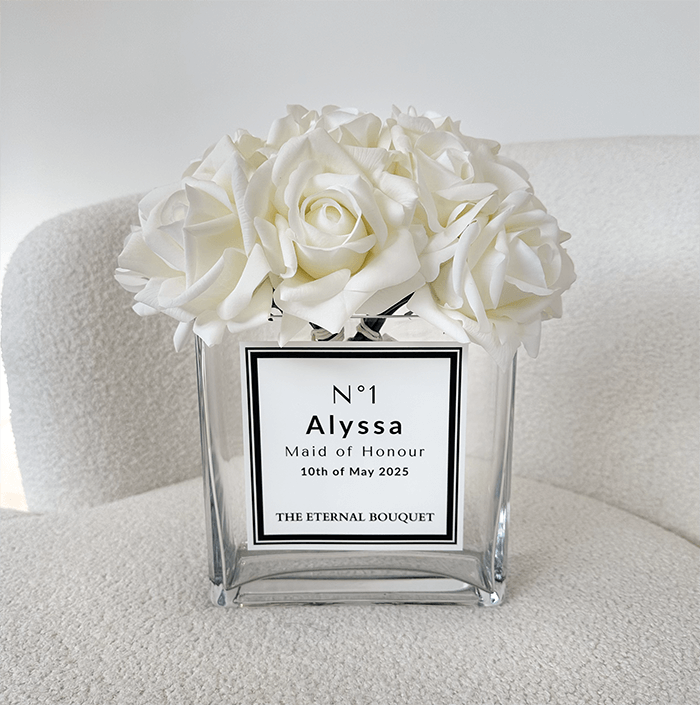 A beautifully arranged bouquet of elegant white roses sits in a clear, square glass vase with a personalized label.The arrangement rests on a plush, cream-colored chair, complementing the minimalist and sophisticated aesthetic of the scene.