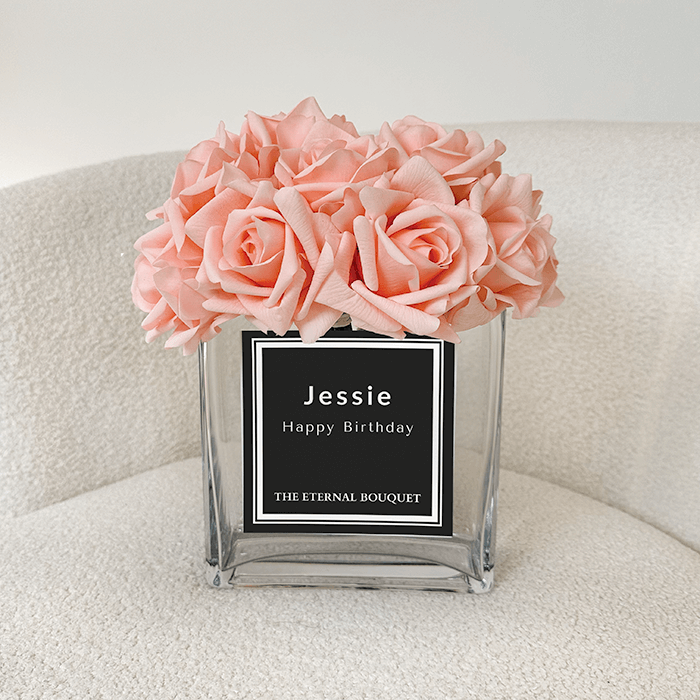 A delicate arrangement of soft pink roses is displayed in a clear, square glass vase with a personalised black label. The bouquet is placed on a plush chair, enhancing the elegant and timeless aesthetic of the presentation.