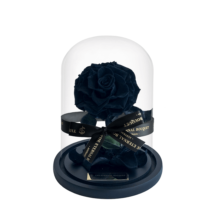A luxurious black everlasting rose elegantly displayed under a glass dome, tied with a sleek black ribbon featuring The Eternal Bouquet’s signature gold branding. Set on a matte black base adorned with scattered petals, this striking arrangement makes a bold, timeless gift for anniversaries, romantic gestures, or sophisticated home décor.