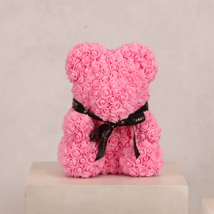 A handcrafted teddy bear made entirely from soft pink everlasting roses, tied with a black ribbon that reads 'The Eternal Bouquet.' Displayed on a pedestal, this adorable floral arrangement makes a timeless and thoughtful gift for any special occasion.