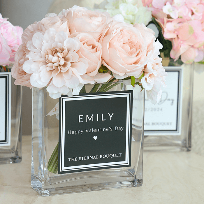 A beautiful arrangement of soft blush pink roses and white dahlias displayed in a clear, square glass vase. The vase features a sleek black and white personalized label, and 