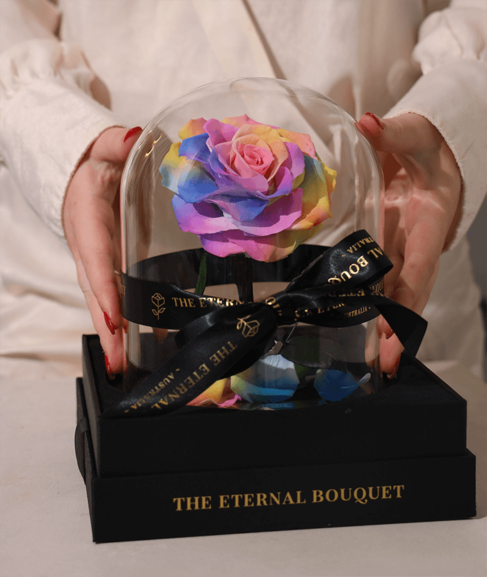 A person holding an everlasting rainbow rose encased in a clear glass dome, elegantly tied with a black ribbon featuring 'The Eternal Bouquet' branding. The dome sits atop a luxurious black gift box with gold lettering that reads 'The Eternal Bouquet. 
