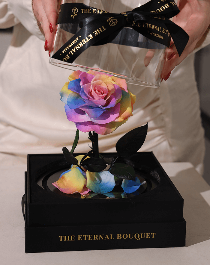 A person lifting the clear glass dome cover, tied with a black ribbon that reads 'The Eternal Bouquet,' to reveal an everlasting rainbow rose arrangement. The vibrant multicolored rose sits on a black round base, surrounded by scattered petals, all displayed on an elegant black gift box labeled 'The Eternal Bouquet' in gold lettering.