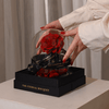 Hands carefully placing an everlasting red rose encased in a glass dome onto a black gift box with gold "The Eternal Bouquet" branding—an elegant and thoughtful luxury gift perfect for romantic occasions.