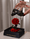 A close-up of a hand gently lifting the glass dome off a beautiful everlasting red rose, set on a sleek black base adorned with scattered petals and elegant gold branding that reads "The Eternal Bouquet." The rose is vibrant and full, symbolizing everlasting love and devotion, making this luxury arrangement a perfect romantic gift for anniversaries, Valentine's Day, or meaningful milestones.