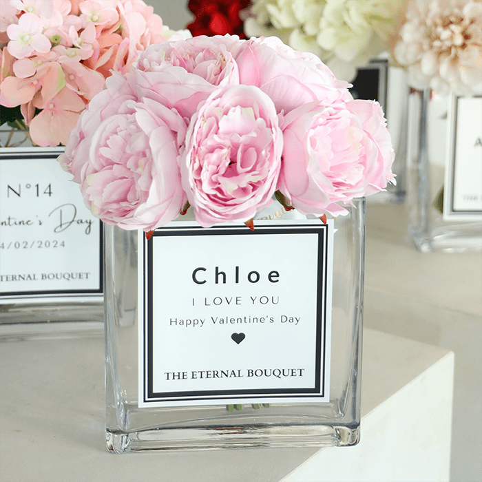 A luxurious floral arrangement from The Eternal Bouquet featuring soft pink peonies displayed in a clear, square glass vase. The vase is personalized with a sleek white label. Surrounding the main arrangement are additional personalized bouquets, creating an elegant and romantic setting. Perfect as gifts for your partners, or to commemorate meanignful memories.