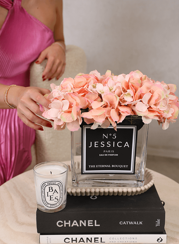 A personalised flower arrangement from The Eternal Bouquet, featuring soft peach everlasting hydrangeas in a clear glass vase labelled 'No. 5 Jessica Paris Eau de Parfum'. The arrangement sits elegantly on a coffee table, styled with Chanel books and a Diptyque candle, creating a luxurious and sophisticated display.