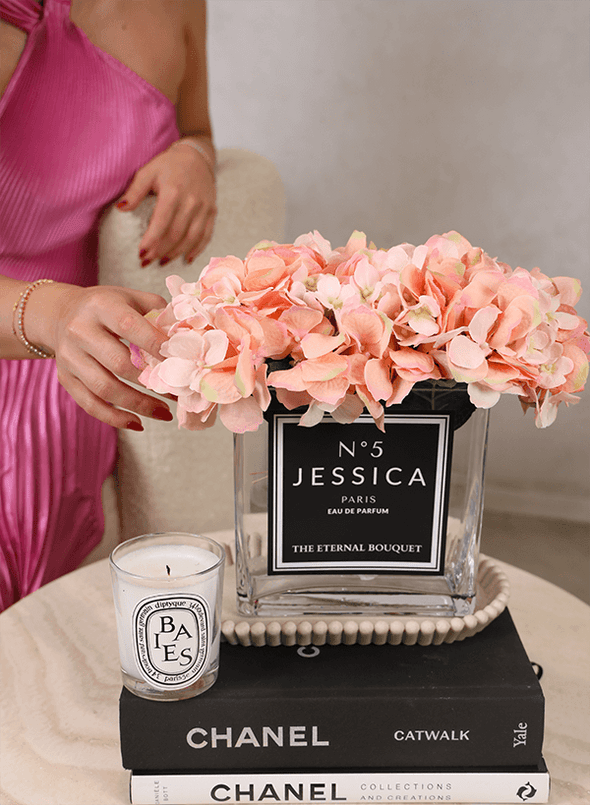 A personalised flower arrangement from The Eternal Bouquet, featuring soft peach everlasting hydrangeas in a clear glass vase labelled 'No. 5 Jessica Paris Eau de Parfum'. The arrangement sits elegantly on a coffee table, styled with Chanel books and a Diptyque candle, creating a luxurious and sophisticated display.