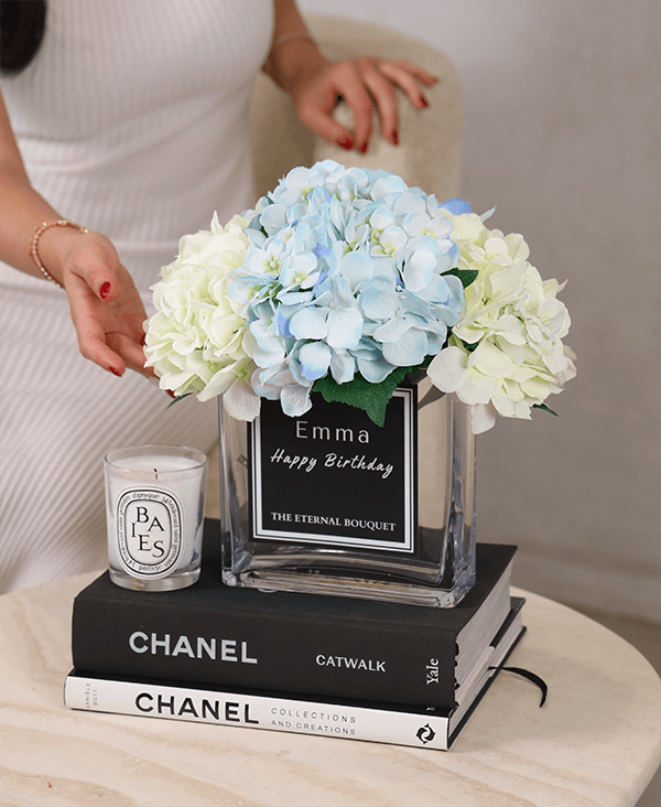 A luxurious arrangement of pale yellow and soft blue hydrangeas displayed in a clear, square glass vase from The Eternal Bouquet. The vase features an elegant black and white personalised label inspired by classic perfume design, reading 