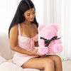 A young woman in a soft pink dress lovingly holds a handcrafted everlasting rose teddy bear from The Eternal Bouquet, made from delicate pink everlasting roses and tied with an elegant black ribbon, the perfect keepsake to express affection in the most charming way.