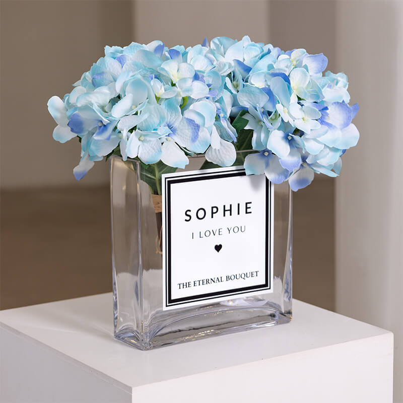 A personalised glass vase from The Eternal Bouquet, featuring delicate preserved blue hydrangeas. The label reads 'Sophie, I Love You,' making it a heartfelt and timeless gift perfect for celebrating love and special moments.