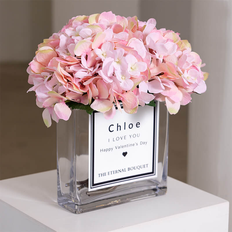 A personalised glass vase from The Eternal Bouquet filled with soft pinkeverlasting hydrangeas. The label reads 'Chloe, I love you, Happy Valentine's Day,' making it a thoughtful and everlasting gift for a special someone.