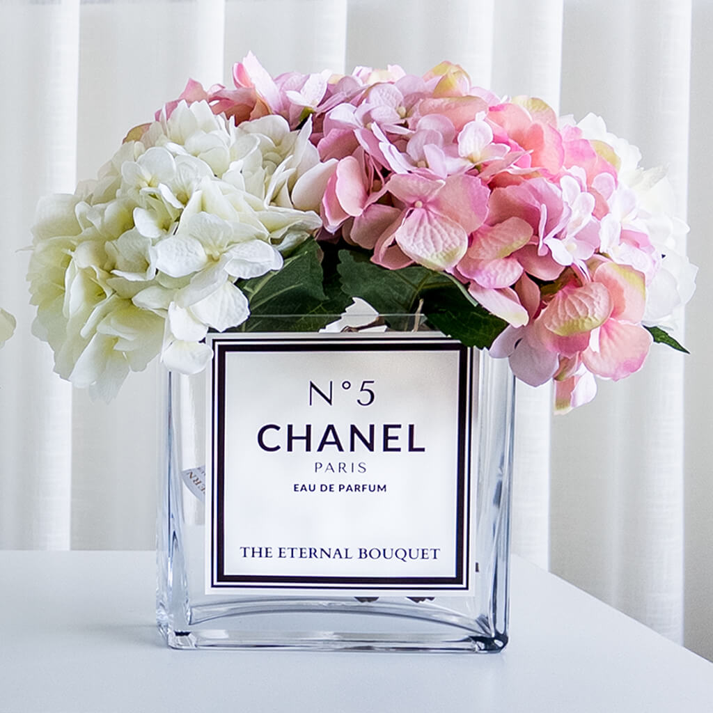 A luxurious floral arrangement by The Eternal Bouquet, featuring elegant white and soft pink hydrangeas displayed in a clear, square glass vase. The vase has a sleek white personalised label with black text, styled like a designer perfume bottle, reading 