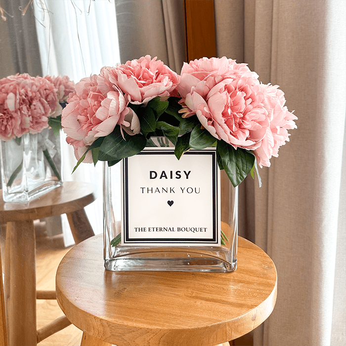 A bouquet of pink peonies with lush green leaves arranged in a clear glass vase with a minimalist label. The vase sits on a round wooden stool in a cozy, modern interior with neutral tones, a wooden mirror, and decorative elements in the background. 