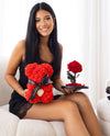 Smiling woman in a black lace dress holding two signature gifts from The Eternal Bouquet, a red rose teddy bear with a pearl heart detail and an everlasting red rose arrangement on a black base, capturing the perfect romantic gesture for Valentine's Day, anniversaries, or special surprises.