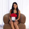 A young woman with long, sleek black hair and a warm smile sits on a cozy brown armchair, holding a luxurious arrangement of vibrant red roses in a clear, square vase. The floral arrangement, from "The Eternal Bouquet," features a personalized label that reads "N°14 Valentine's Day."  The background features soft white curtains, adding a bright and airy feeling to the image.
