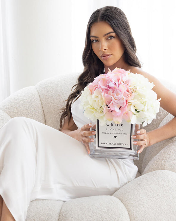 A luxurious floral arrangement by The Eternal Bouquet, featuring elegant white and soft pink hydrangeas displayed in a clear, square glass vase. The vase has a sleek white personalised label with black text, styled like a designer perfume bottle, reading "N°5 CHANEL PARIS EAU DE PARFUM" and "THE ETERNAL BOUQUET" at the bottom. The arrangement sits on a white surface with soft, sheer curtains in the background, creating an elegant and sophisticated presentation.