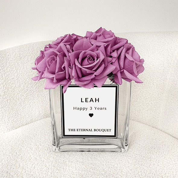 A beautiful arrangement of vibrant purple roses is elegantly displayed in a clear, square glass vase. The bouquet is placed on a soft, cream-colored chair, enhancing its luxurious and timeless appeal.
