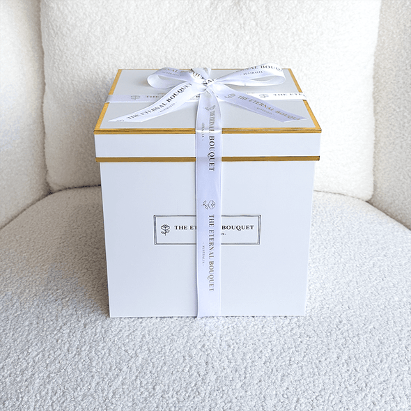 A luxurious white gift box with gold accents and a neatly tied white ribbon, featuring "THE ETERNAL BOUQUET" branding. The elegant packaging sits on a soft, textured surface, exuding a sophisticated and premium feel. This box is designed to hold the everlasting floral arrangements from The Eternal Bouquet, making it a perfect gift for special occasions.