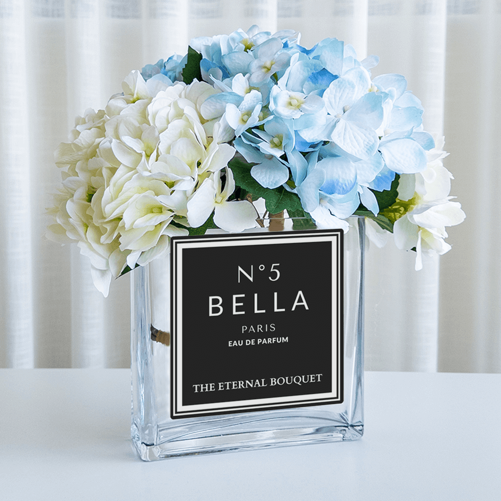 A luxurious arrangement of pale yellow and soft blue hydrangeas displayed in a clear, square glass vase from The Eternal Bouquet. The vase features an elegant black and white personalised label inspired by classic perfume design, reading 