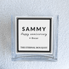 A close-up view of a personalized message label on a clear, square glass vase from The Eternal Bouquet. The label features a minimalist black and white design. The vase sits on a soft, textured white surface, emphasizing its elegant and timeless aesthetic.