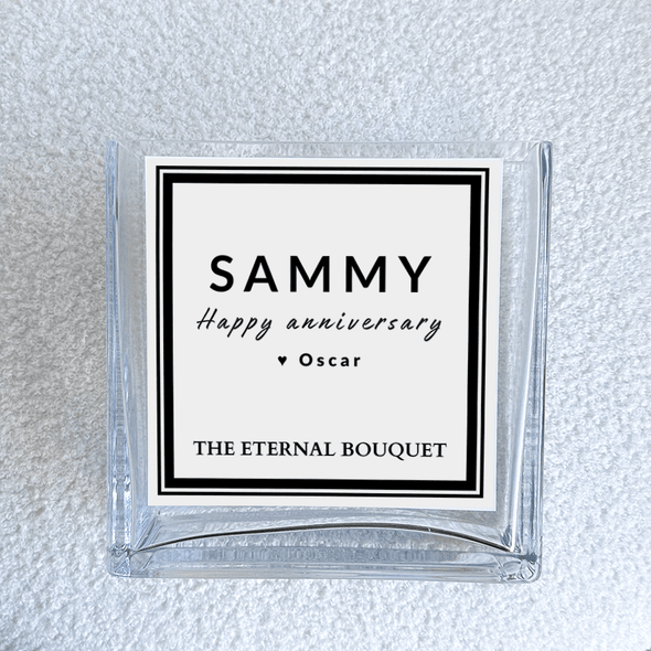 A close-up view of a personalized message label on a clear, square glass vase from The Eternal Bouquet. The label features a minimalist black and white design. The vase sits on a soft, textured white surface, emphasizing its elegant and timeless aesthetic.