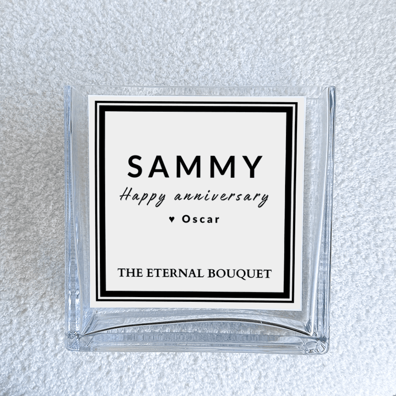 A close-up view of a personalized message label on a clear, square glass vase from The Eternal Bouquet. The label features a minimalist black and white design. The vase sits on a soft, textured white surface, emphasizing its elegant and timeless aesthetic.