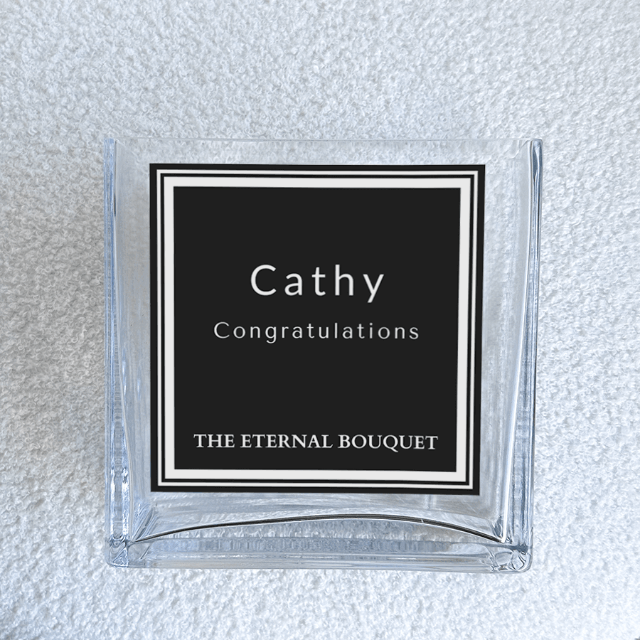 A close-up of a personalized label on a clear, square glass vase from The Eternal Bouquet. The vase is placed on a soft, textured white surface, highlighting its modern and sophisticated design