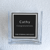 A close-up of a personalized label on a clear, square glass vase from The Eternal Bouquet. The vase is placed on a soft, textured white surface, highlighting its modern and sophisticated design