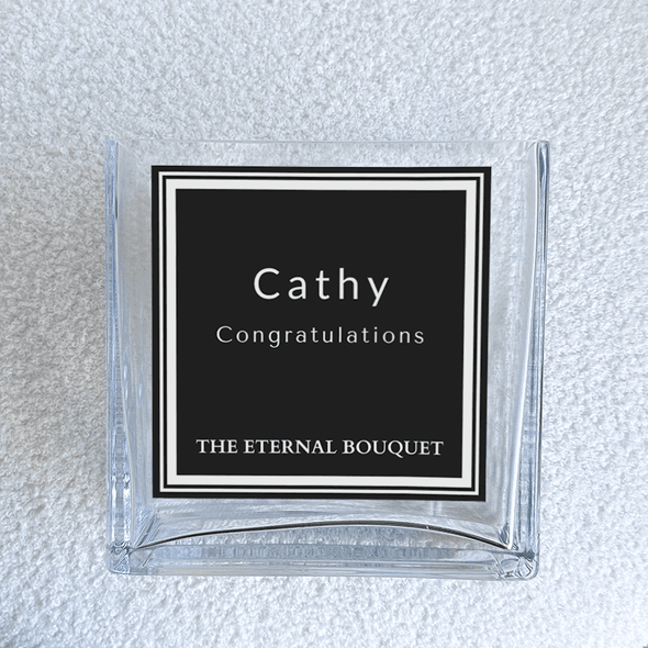 A close-up of a personalized label on a clear, square glass vase from The Eternal Bouquet. The vase is placed on a soft, textured white surface, highlighting its modern and sophisticated design