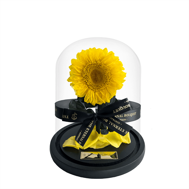 A stunning everlasting sunflower encased in a sleek glass dome, tied with an elegant black ribbon featuring 'The Eternal Bouquet' in gold lettering. Resting on a sophisticated black base, this timeless arrangement captures the warmth and joy of sunflowers, symbolizing happiness and positivity. A perfect gift for brightening someone’s day, celebrating special moments, or adding a cheerful touch to any space. 