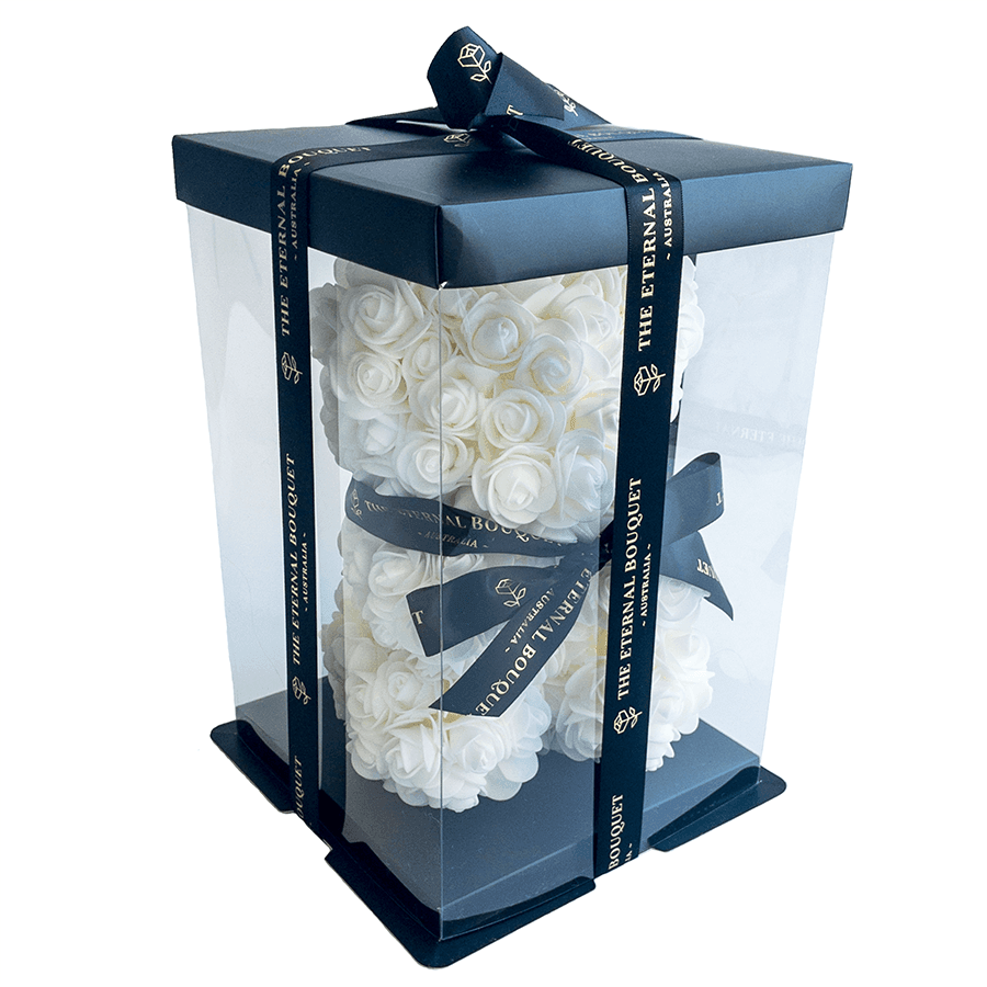A sophisticated everlasting white rose teddy bear enclosed in a clear display gift box, wrapped with sleek black ribbon featuring The Eternal Bouquet’s gold-accented branding. The delicate ivory roses and premium packaging make it a timeless and elegant present—perfect for weddings, anniversaries, or luxurious décor.